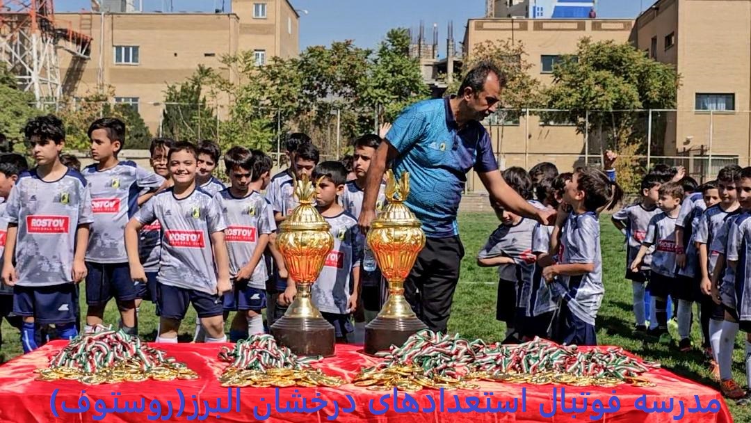 تصویر جشن قهرمانی با میزی پر از مدال در زمین چمن طبیعی با بازیکنان نونهال با لباس طوسی که از مربی خود با لباس آبی مدال می گیرند