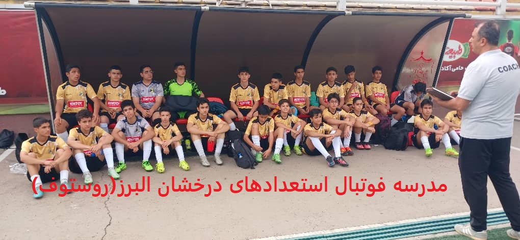 تصویر نشان‌دهنده تیم نوجوانان مدرسه فوتبال ایران روستوف با لباس زرد و مشکی و لوگوی "ROSTOV" نشسته روی نیمکت کنار زمین چمن و گوش دادن به مربی خود با لباس طوسی